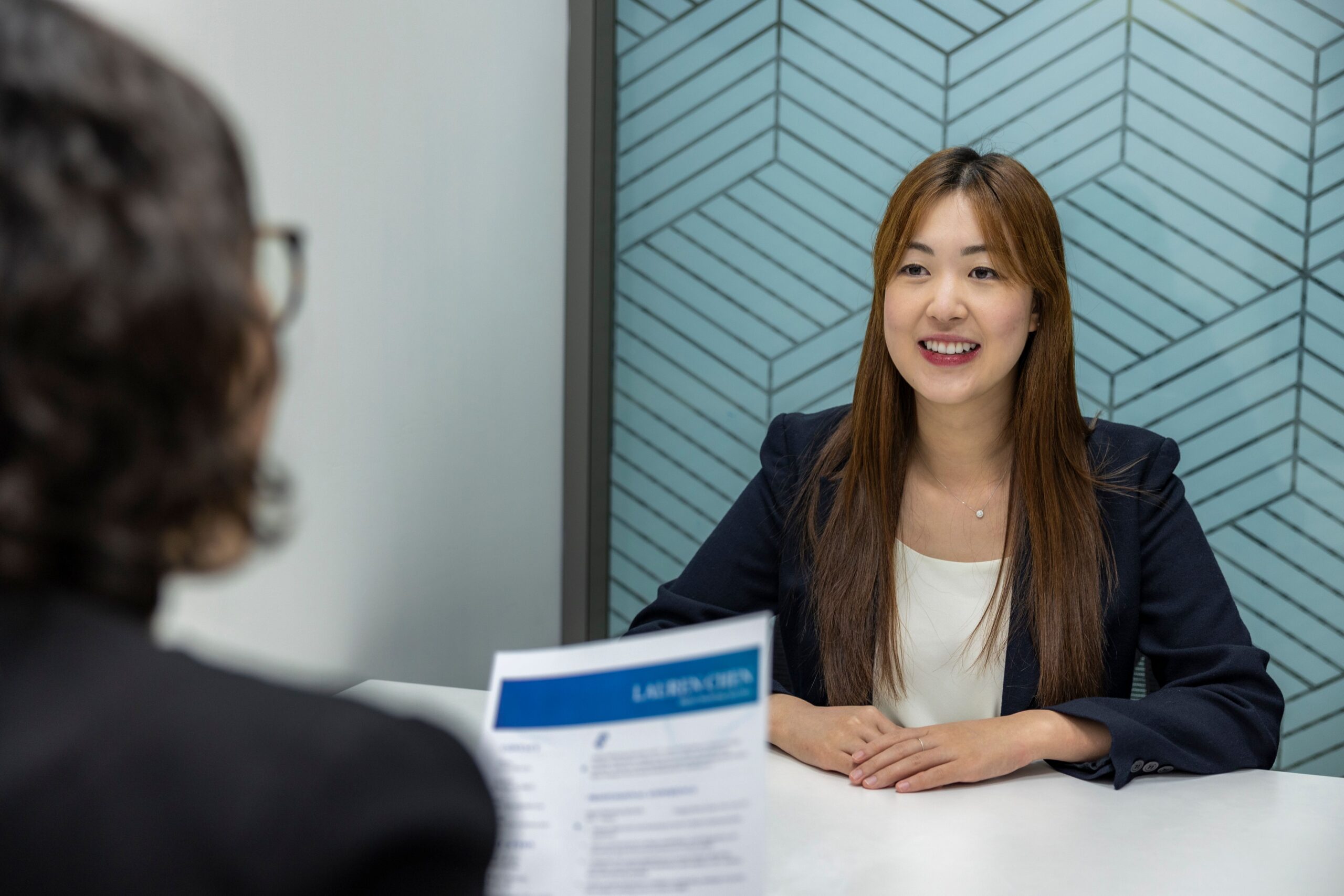 Woman in blazer smiling at interview across desk, interviewer holding resume